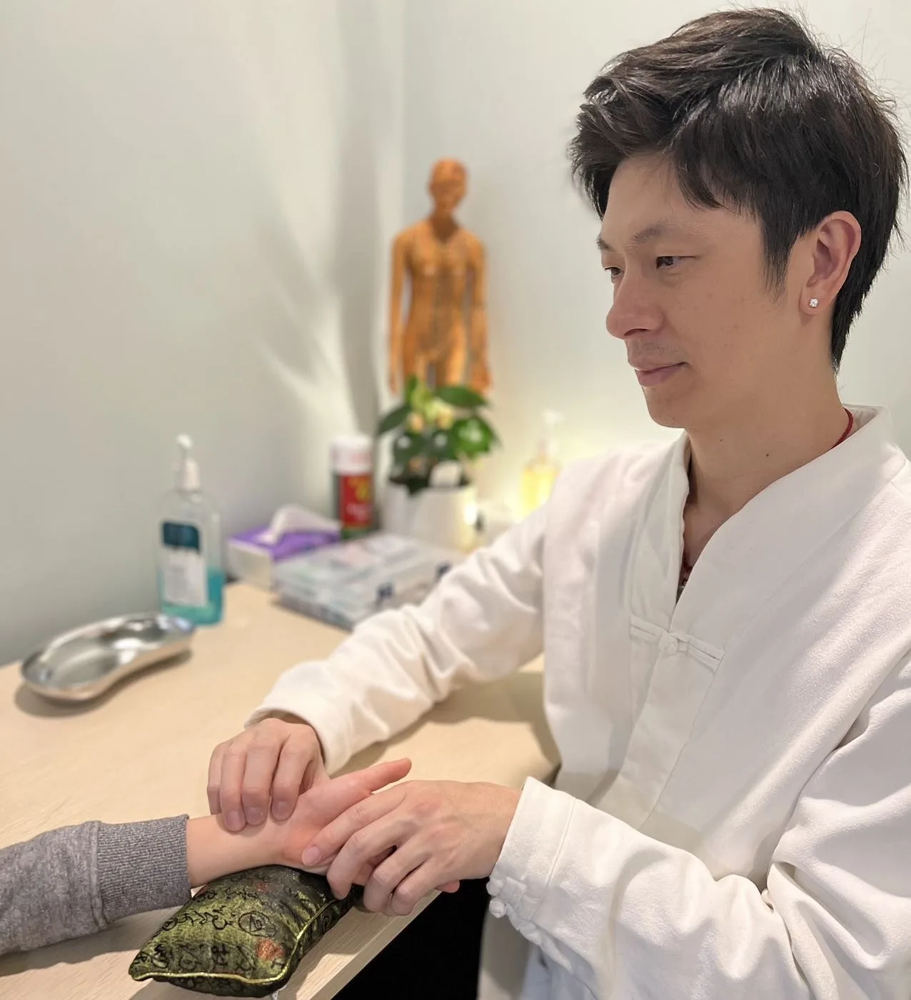 Ken Hui in a white clinical tunic, taking a patient's wrist pulse with a traditional acupoint model behind him