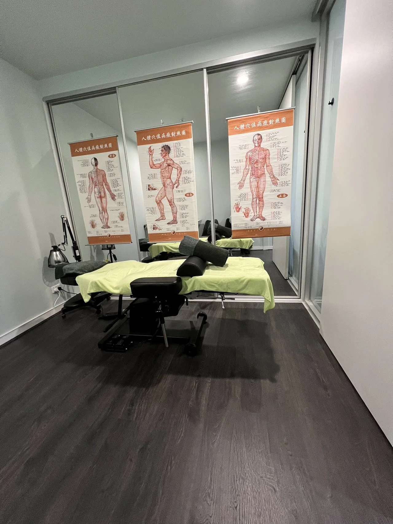 One Acupuncture treatment room with Chinese meridian point charts on a mirrored wardrobe, green-draped tables, and dark timber floor
