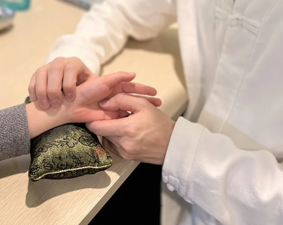 Ken's hands taking a patient's pulse on a green brocade wrist pillow — traditional Chinese pulse diagnosis